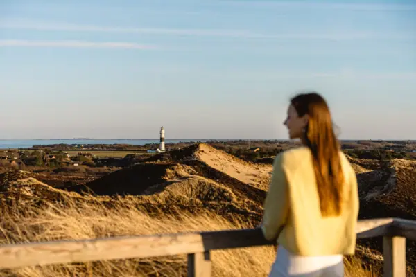 Sylt Inseltour Eine Frau steht an einem Zaun und blickt auf einen Leuchtturm und Dünen.