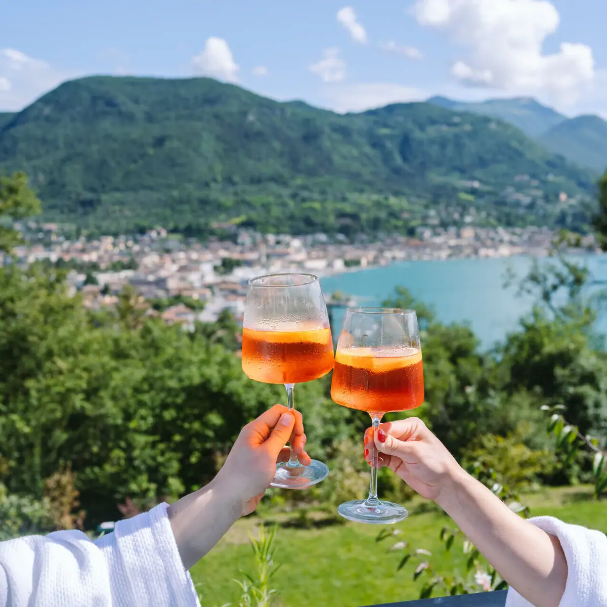 Aperol am Gardasee Zwei Hände halten Weingläser mit Aperol Spritz vor dem Gardasee.