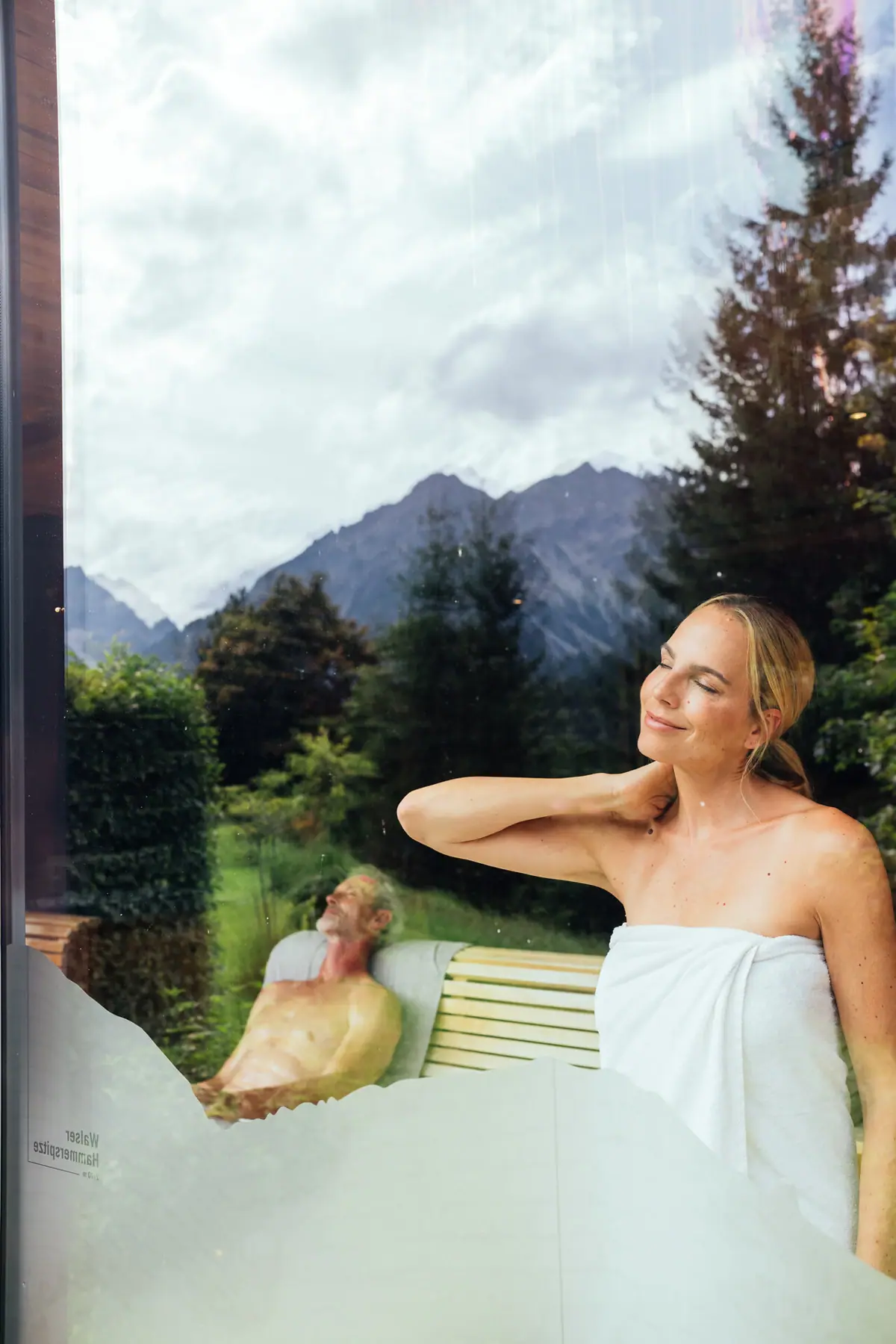 Panoramasauna A-ROSA Ifen Hotel Kleinwalsertal Eine Frau schaut aus dem Fenster einer Panoramasauna, in dem sich der Ausblick spiegelt.