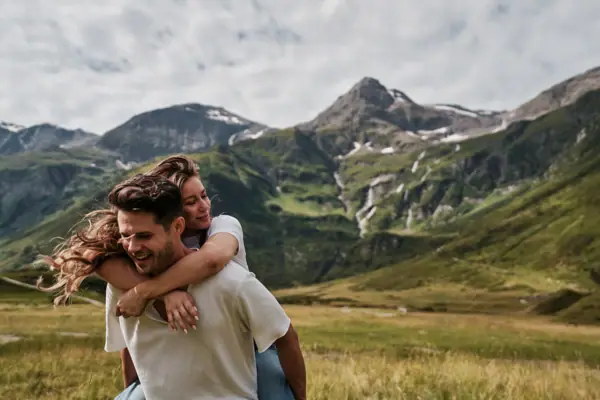 Sommer in Bad Gastein Ein Mann trägt eine Frau auf dem Rücken in einem grasbewachsenen Feld mit Bergen im Hintergrund.