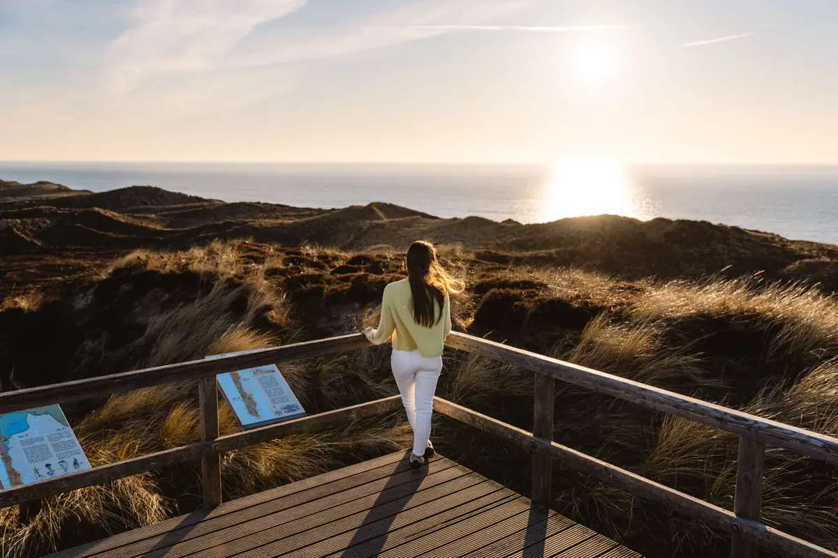 Dünen Sylt Eine Frau steht auf einer Holzbrücke und blickt auf die Wanderdünen und die Nordsee auf Sylt.