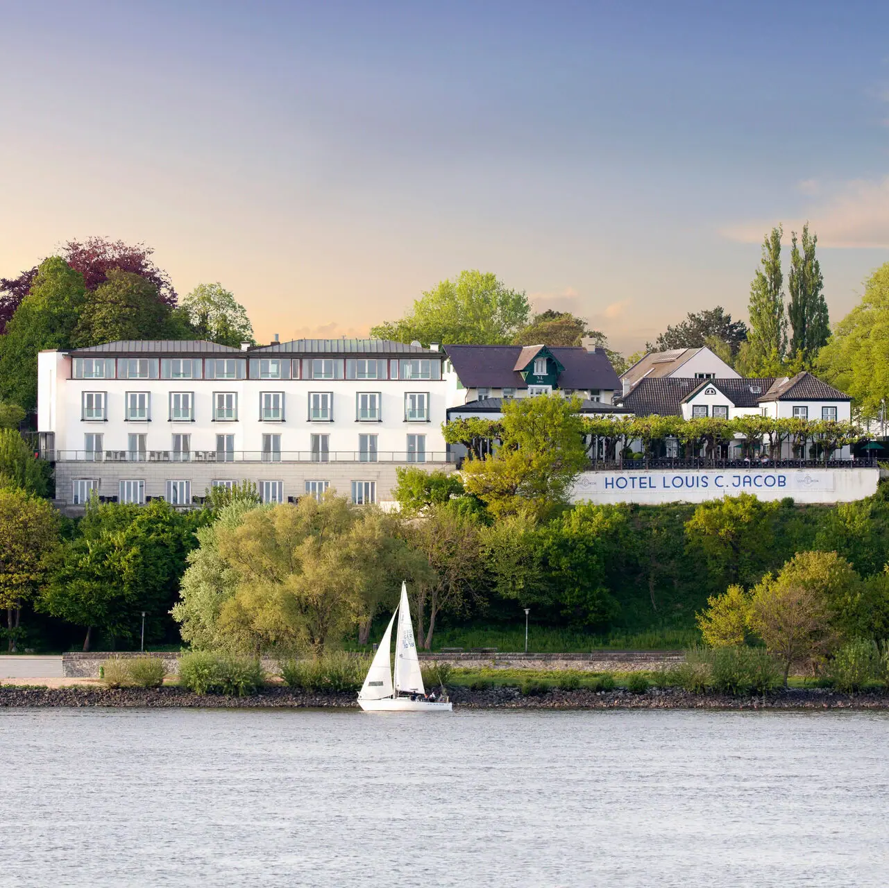 Das Louis C. Jacob Hotel Hamburg von außen mit einem Segelboot davor auf der Elbe.