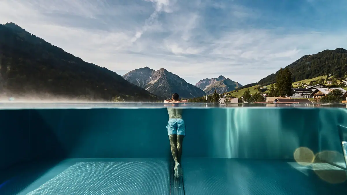 Ausblick Ifen Hotel Kleinwalsertal Ein Mann im Outdoor-Pool vom A-ROSA Ifen Hotel Kleinwalsertal mit Bergen im Hintergrund.
