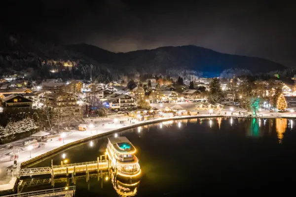 Pendelschiff Tegernsee Weihnachtsmarkt Eine Stadt bei Nacht mit Lichtern, die sich im Wasser spiegeln.