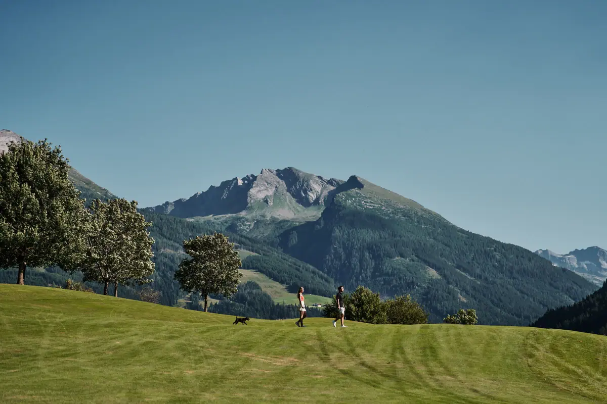 Wandern Menschen wandern auf einem grasbewachsenen Hügel mit Bäumen und einem Berg im Hintergrund.